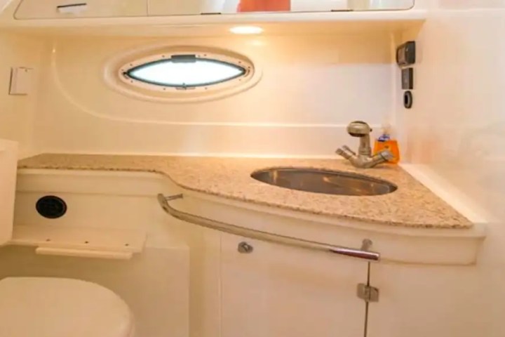 Small boat bathroom with a circular porthole, sink, and toilet; curved wooden countertop with a faucet.