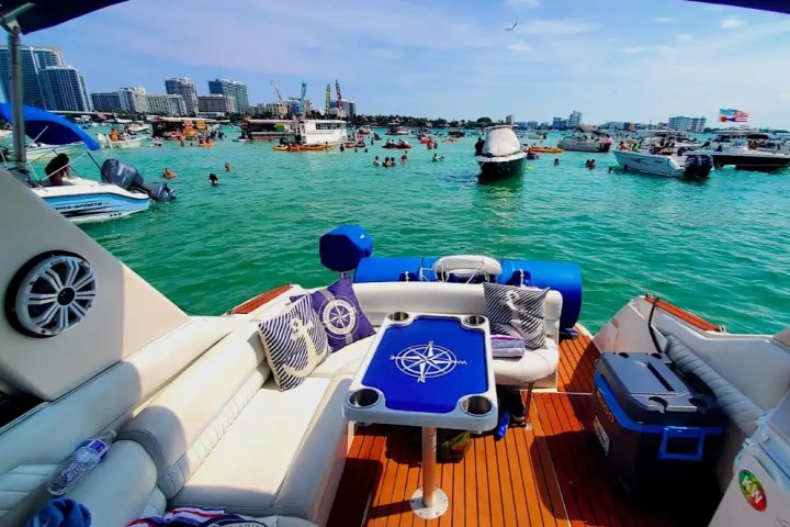 View from a boat with seating, looking at other boats and swimmers in turquoise water near city buildings.