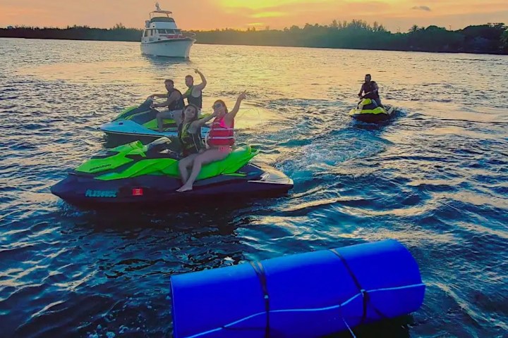 People riding jet skis on a lake with sunset and a boat in the background.