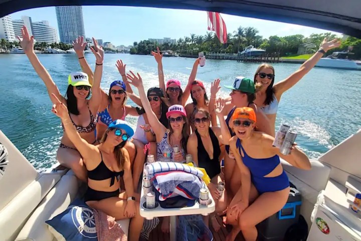 Group of women in swimsuits on a boat, wearing hats and holding drinks, with arms raised and waterway in background.