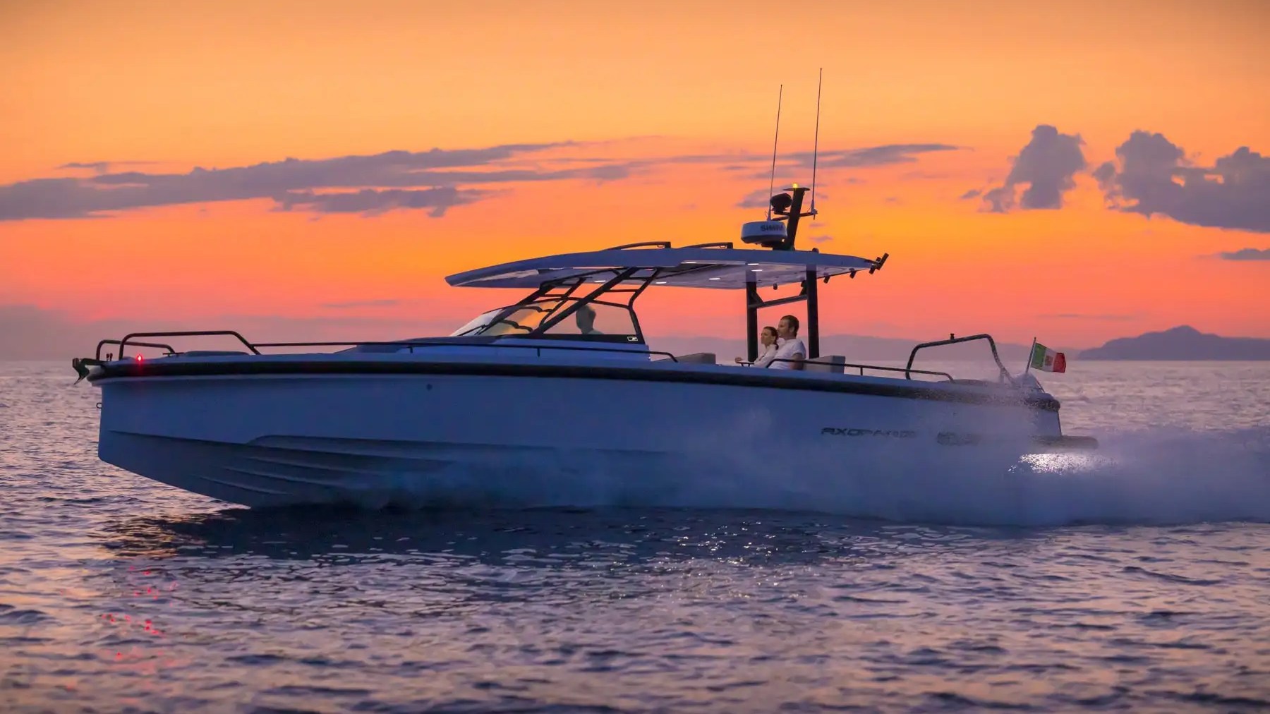 Boat cruising on ocean at sunset, with a couple onboard and Italian flag waving at the back.