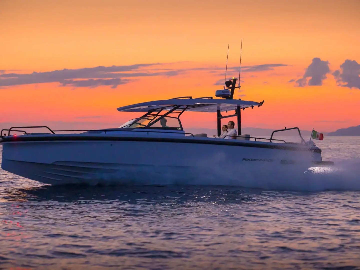Boat cruising on ocean at sunset, with a couple onboard and Italian flag waving at the back.