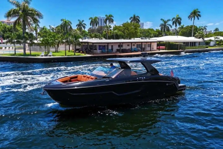 Luxury boat on blue water near palm-lined shore and modern house under clear sky.