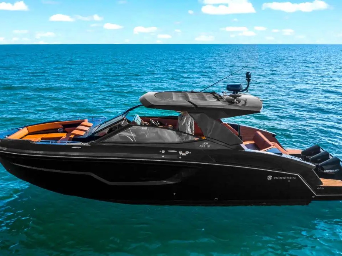 This 38 foot Cruisers Boat Rental is shown from an aerial side profile cruising Gulf Coast waters near Naples, featuring a sleek black hull, hardtop design, open bow lounge, and triple outboard engines.