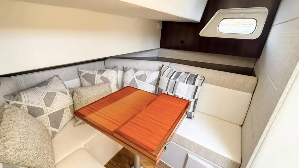 Cozy boat cabin with white cushioned seats, wooden table, pillows, and a small window.