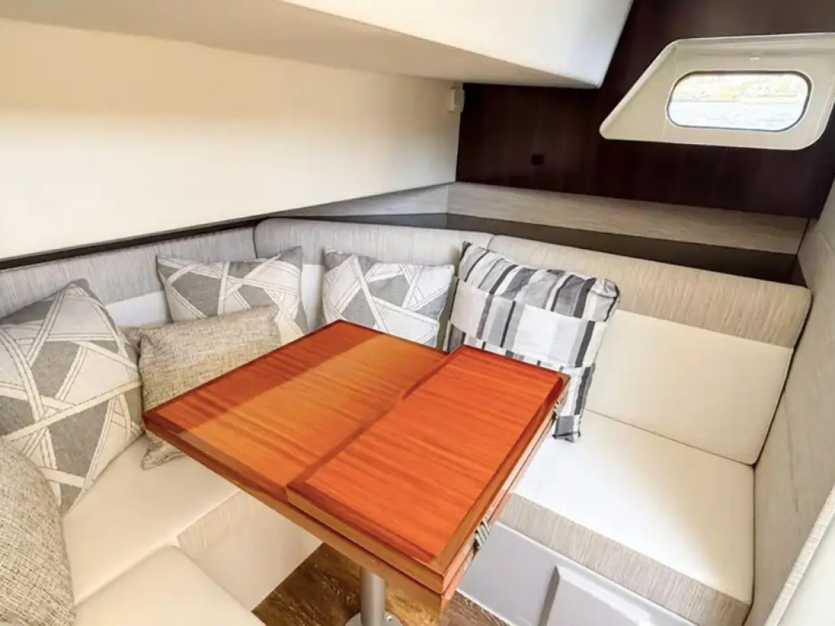 This 38 foot Cruisers Boat Rental features a private cabin lounge with wraparound seating, wood dining table, decorative cushions, and natural light from the side window while cruising Naples waters.