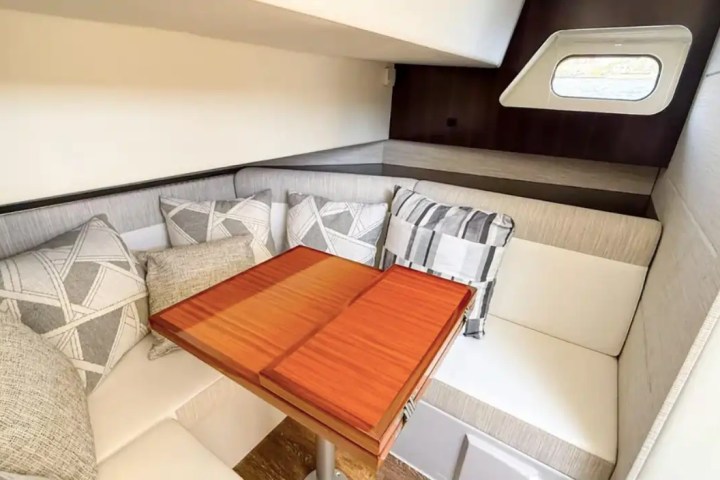 Cozy boat cabin with white cushioned seats, wooden table, pillows, and a small window.