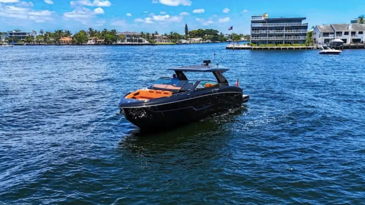 Sleek black yacht on blue water with buildings and trees in the background under a clear sky.