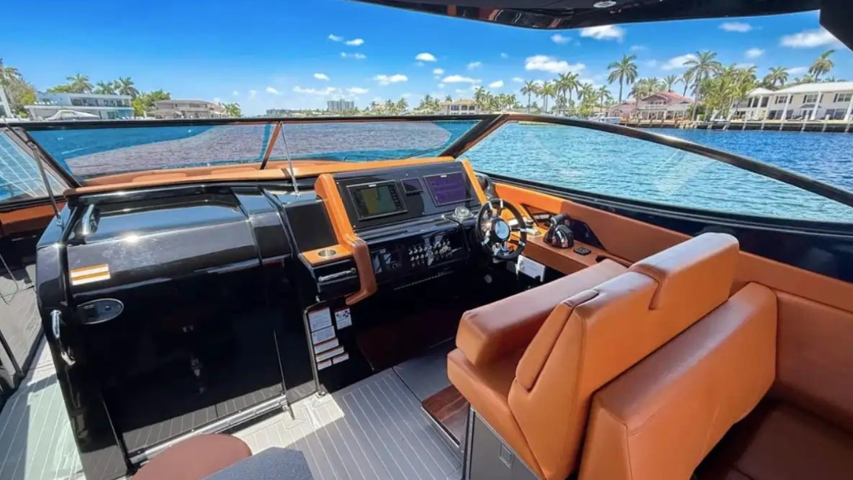 Boat cockpit with brown seats, steering wheel, and controls, overlooking a waterfront with palm trees.