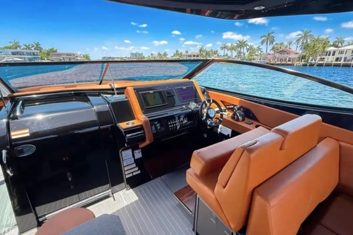 Boat cockpit with brown seats, steering wheel, and controls, overlooking a waterfront with palm trees.