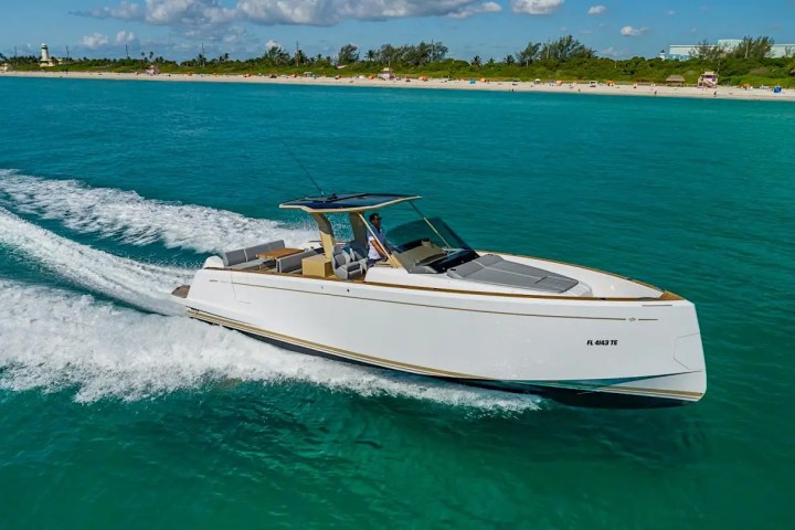 A white yacht cruising on turquoise water near a sandy beach with trees in the background.