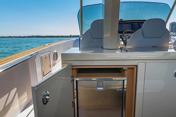 Boat cockpit with open cabinet revealing a mini fridge, overlooking water.
