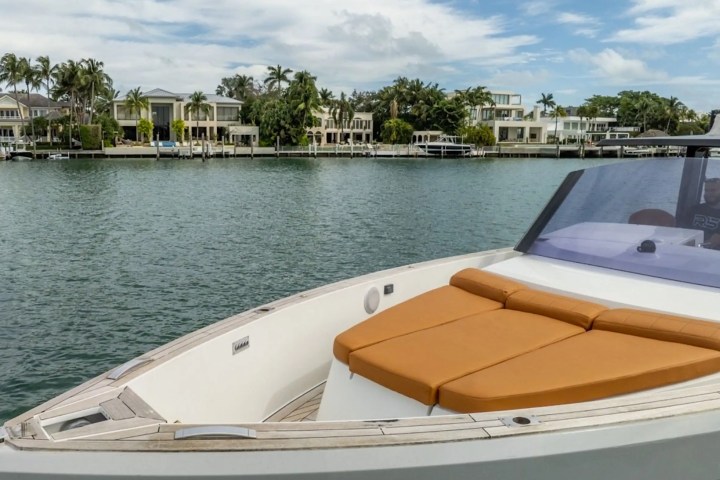 Boat with tan seating cruising near waterfront homes and palm trees under blue sky.