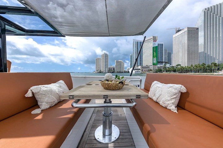 Yacht deck with beige cushions and pillows, table, city skyline and water view under cloudy sky.