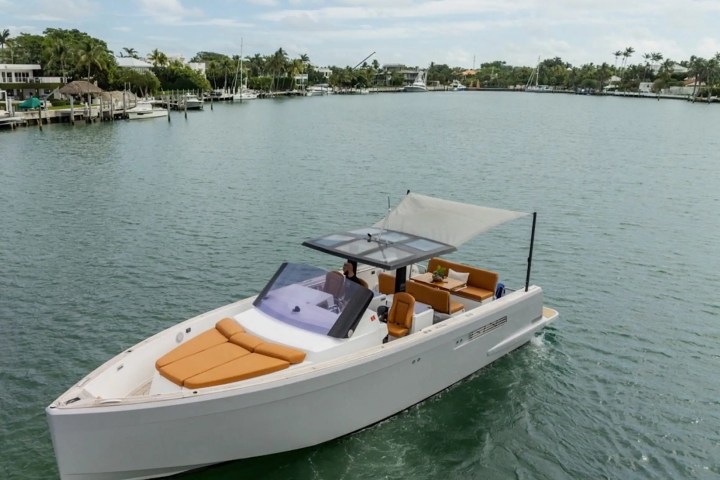 Modern white boat with orange seating on a calm lake, surrounded by green trees and houses.
