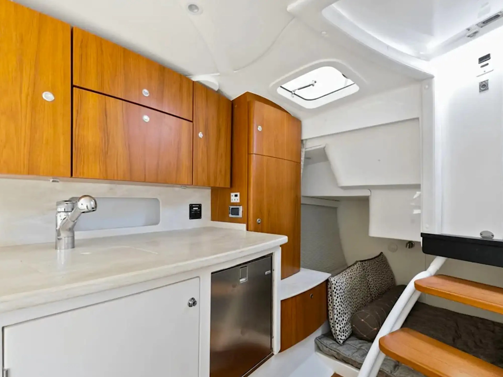 Compact boat kitchenette with wooden cabinets, white countertops, and stairs leading to a cozy seating area.