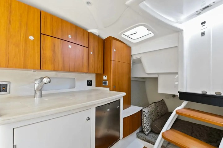 Compact boat kitchenette with wooden cabinets, white countertops, and stairs leading to a cozy seating area.
