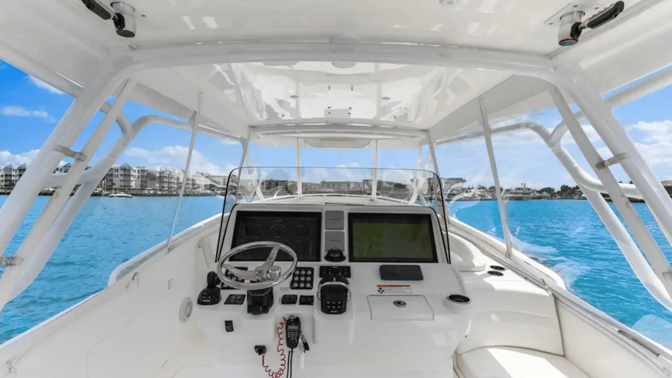 White boat cockpit with steering wheel and control panels, overlooking blue water and distant shoreline.