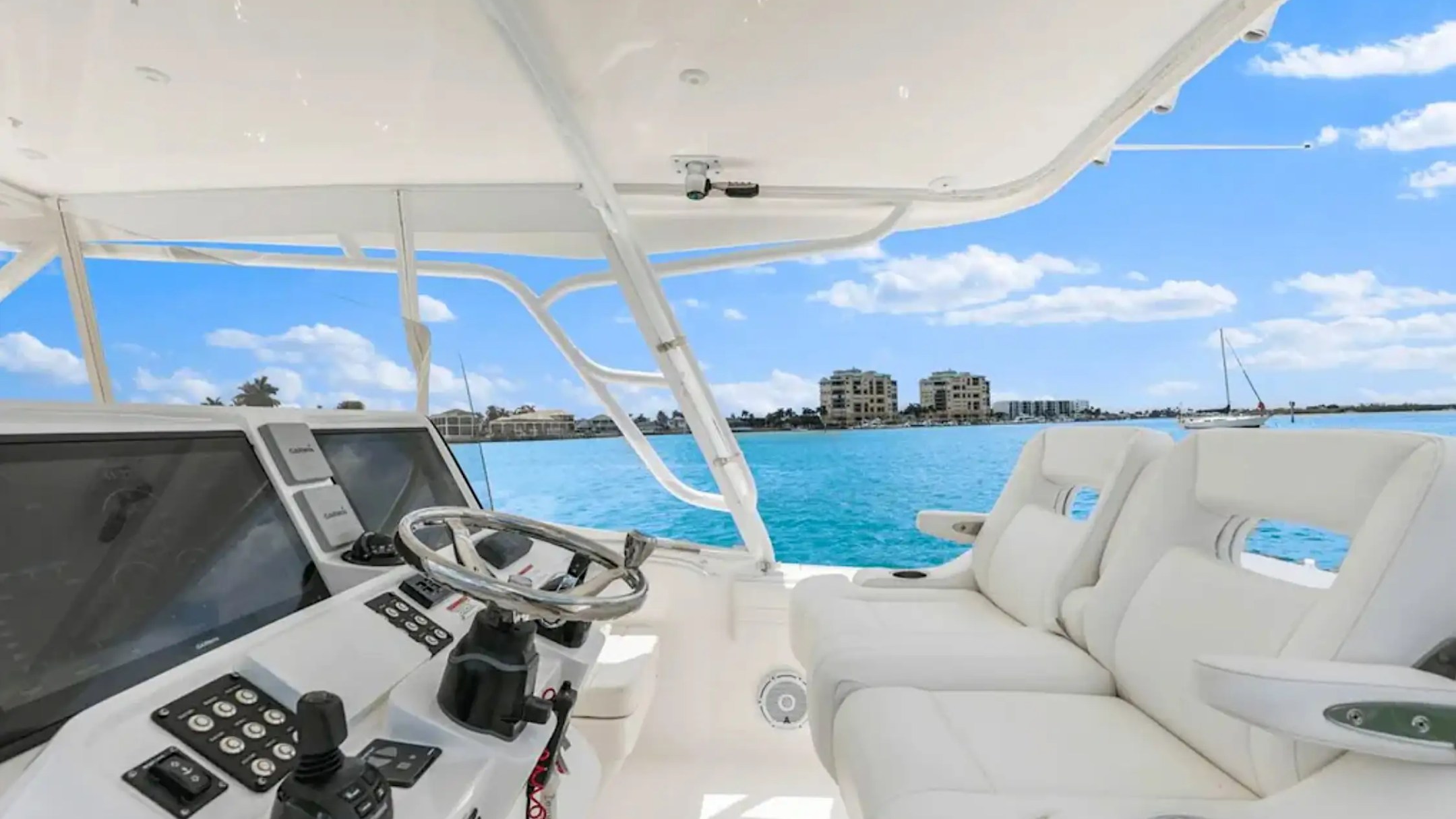 Boat helm with controls and steering wheel, white seats, ocean view, and distant buildings under blue sky.