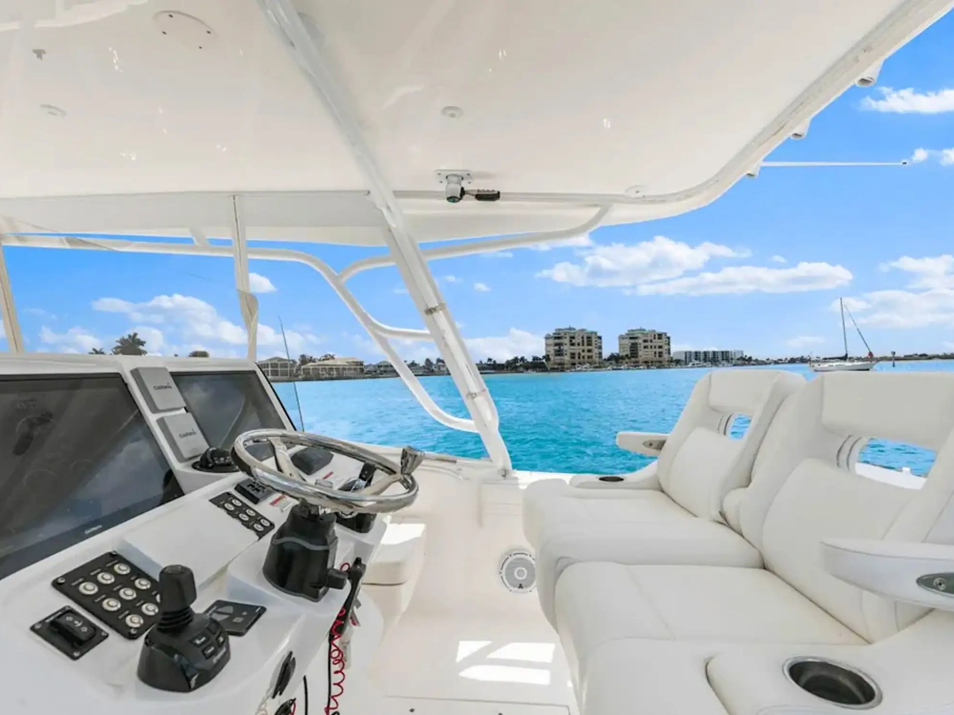 Boat helm with controls and steering wheel, white seats, ocean view, and distant buildings under blue sky.