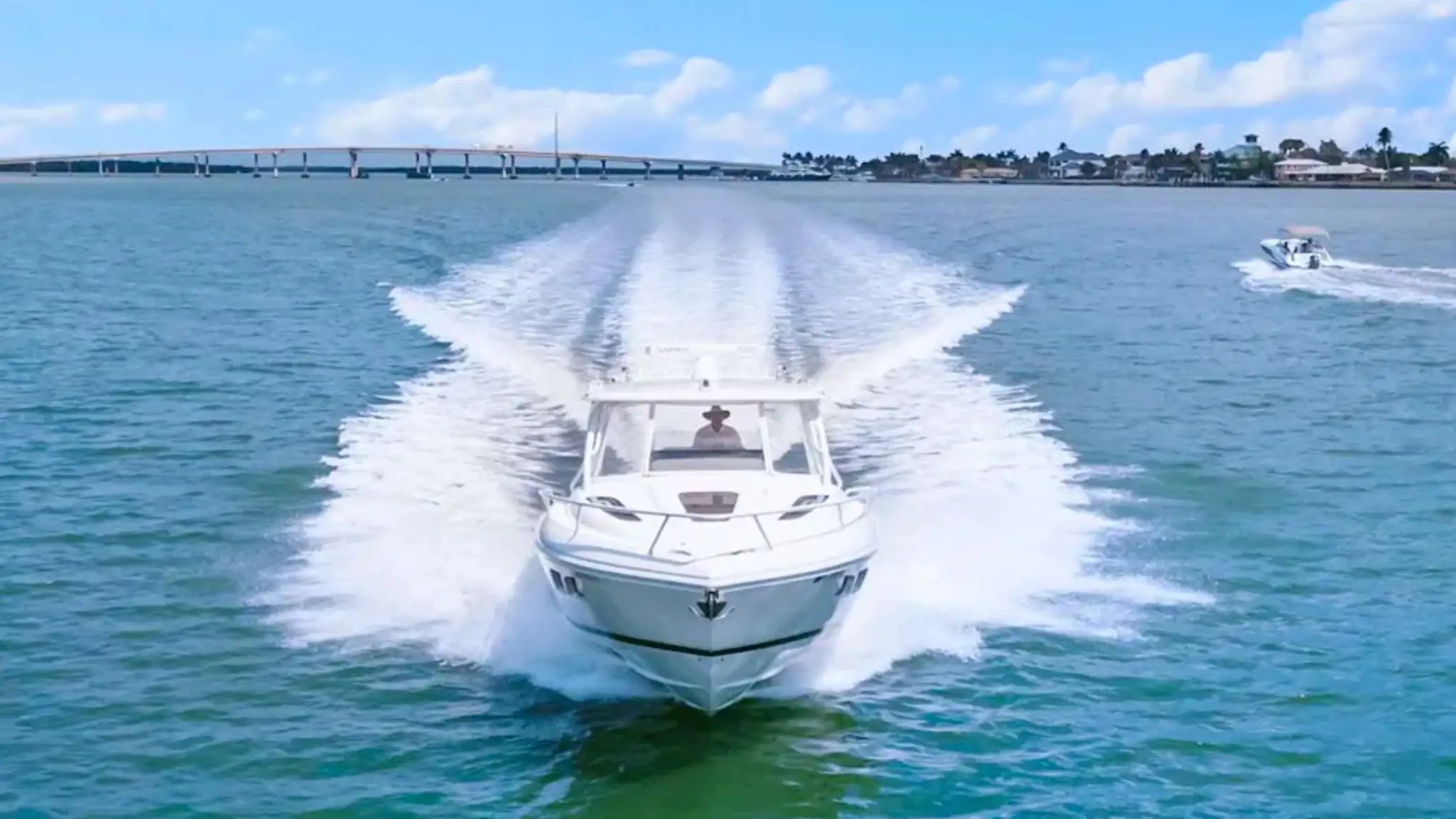 Speedboat moving quickly on blue water with bridge and other boat in background.