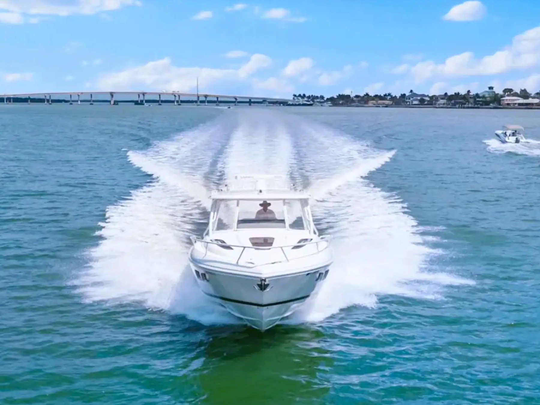 Speedboat moving quickly on blue water with bridge and other boat in background.