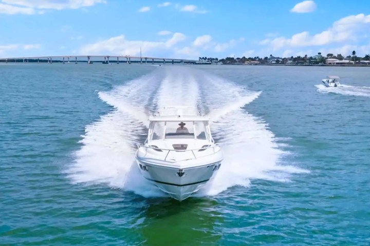 Speedboat moving quickly on blue water with bridge and other boat in background.