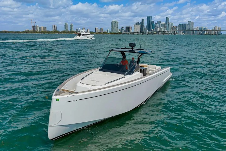 A sleek white yacht on blue water with a city skyline in the background and partly cloudy sky.