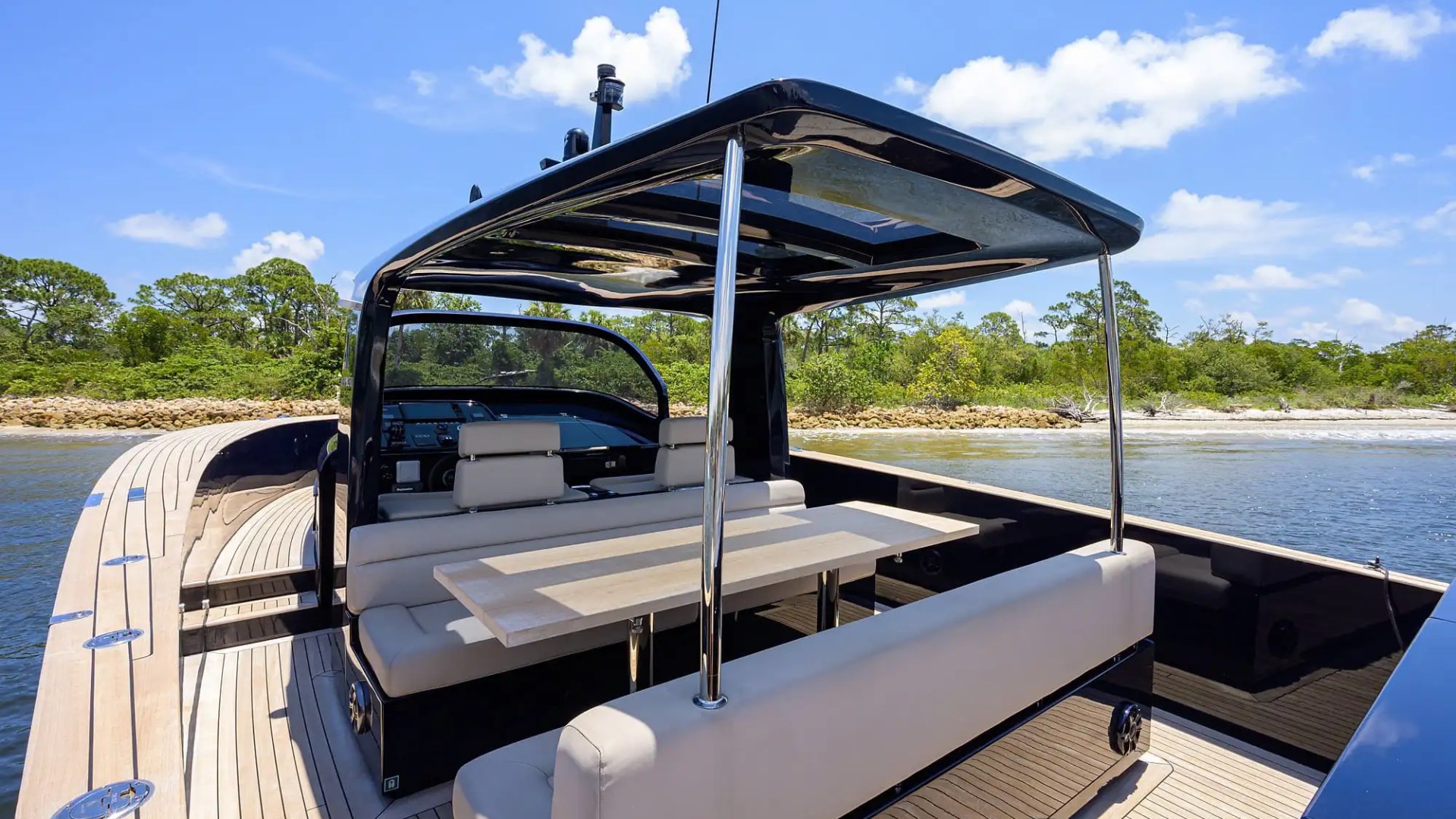 Luxury yacht interior with seating and table, set near a shoreline with trees under a blue sky.