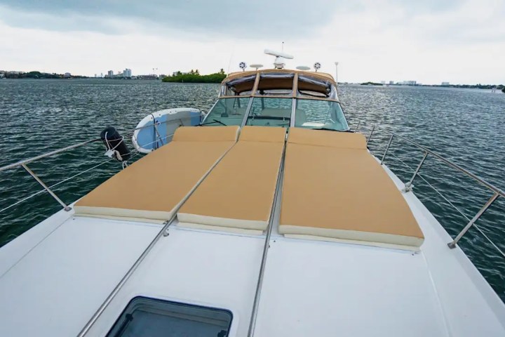 View from bow of a yacht with tan cushions, anchored in a calm body of water with buildings in the distance.