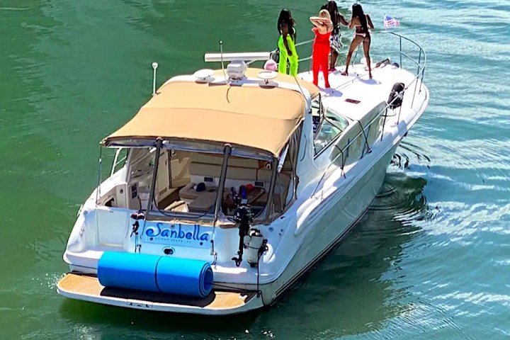 Boat with four people on deck in bright outfits, cruising on water.