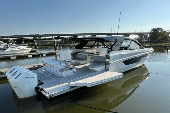 Modern boat docked at a marina with other boats and a bridge in the background.