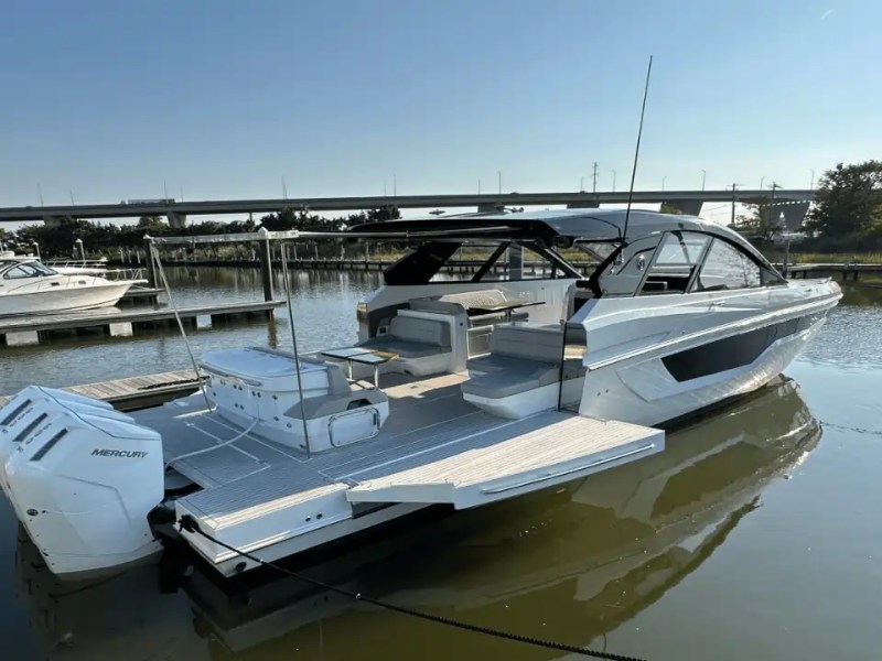 Modern boat docked at a marina with other boats and a bridge in the background.