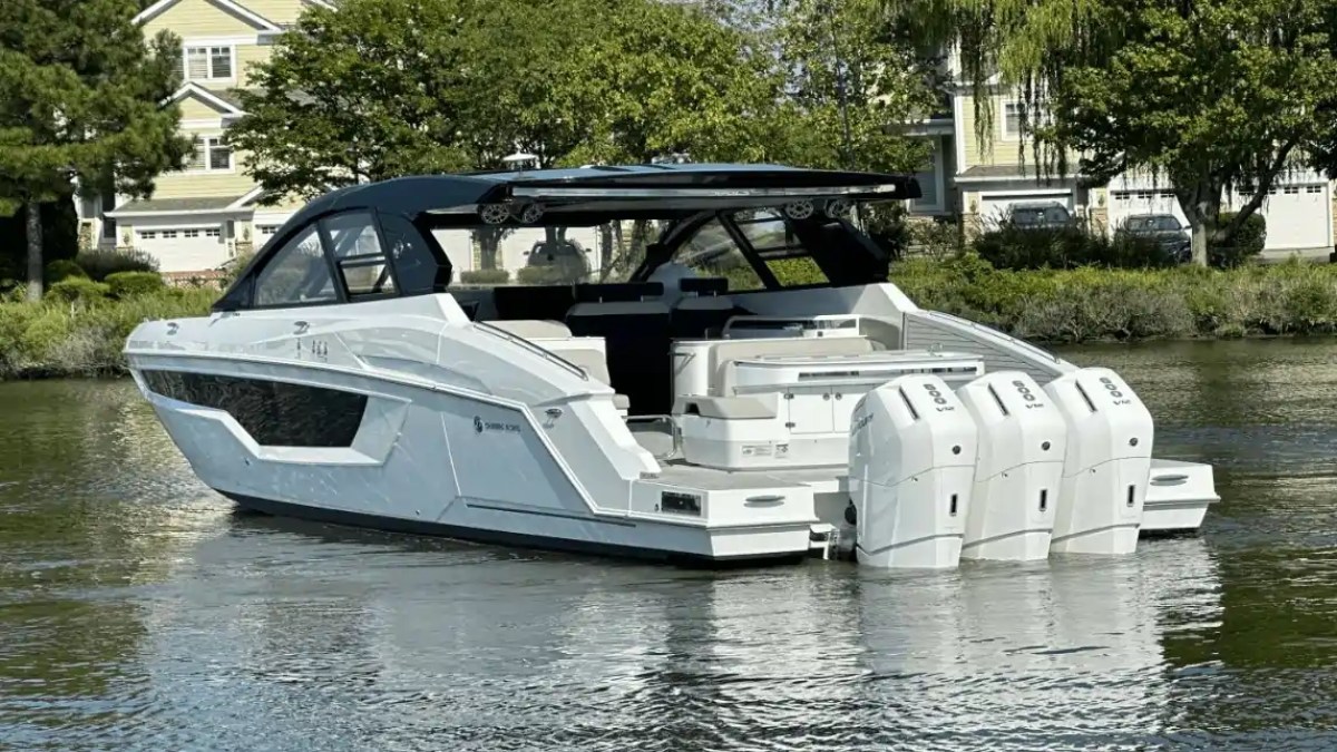 A modern white motorboat with three engines, docked near houses and trees.