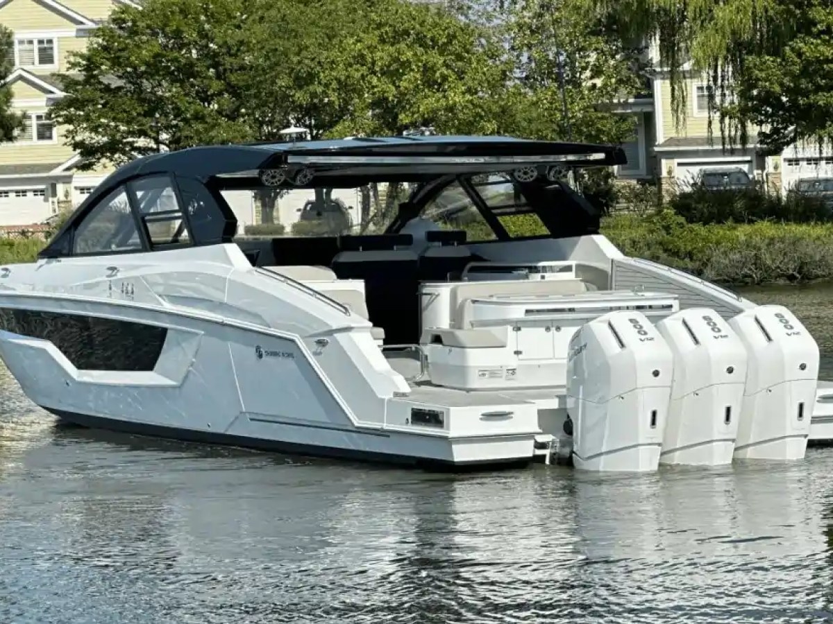 A modern white motorboat with three engines, docked near houses and trees.