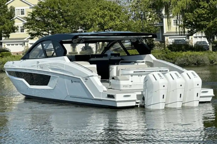 A modern white motorboat with three engines, docked near houses and trees.