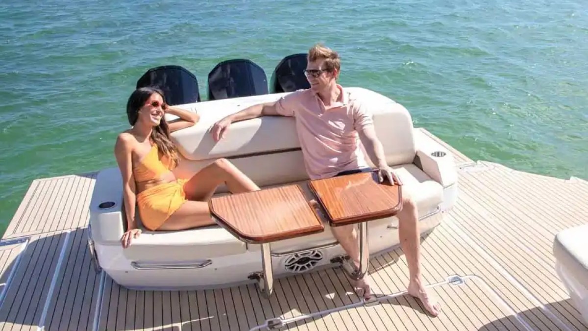 A woman and man relax on a boat deck by the ocean, seated on white cushions with wooden tables.