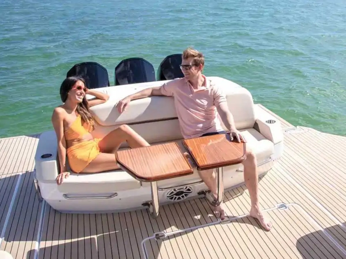 A woman and man relax on a boat deck by the ocean, seated on white cushions with wooden tables.