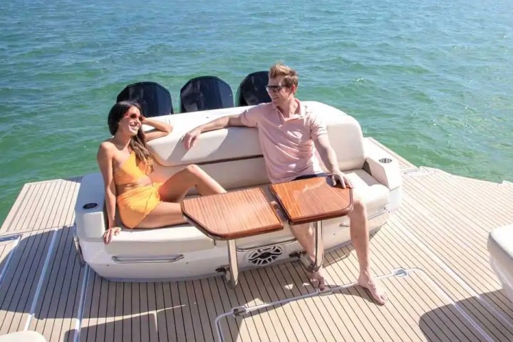 A woman and man relax on a boat deck by the ocean, seated on white cushions with wooden tables.