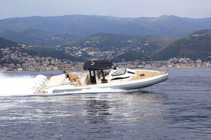 Speedboat with people cruising on a calm sea, mountains and coastal city in the background.