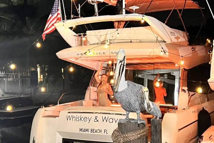 Two people on a yacht at night, with string lights and an American flag, near a decorative pelican figure.
