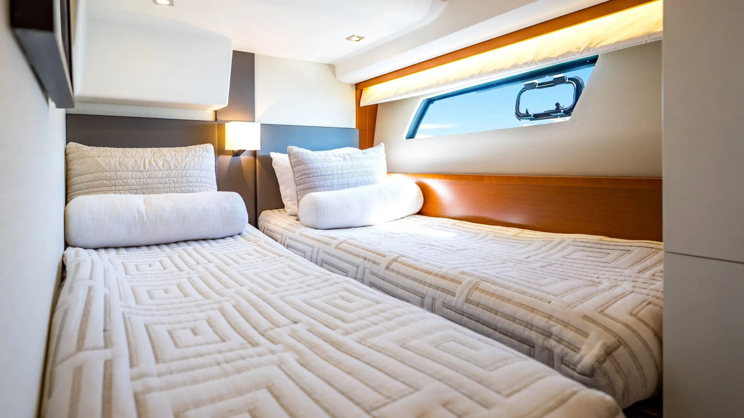 Two twin beds with patterned bedding on a yacht, under a window and accent lighting.