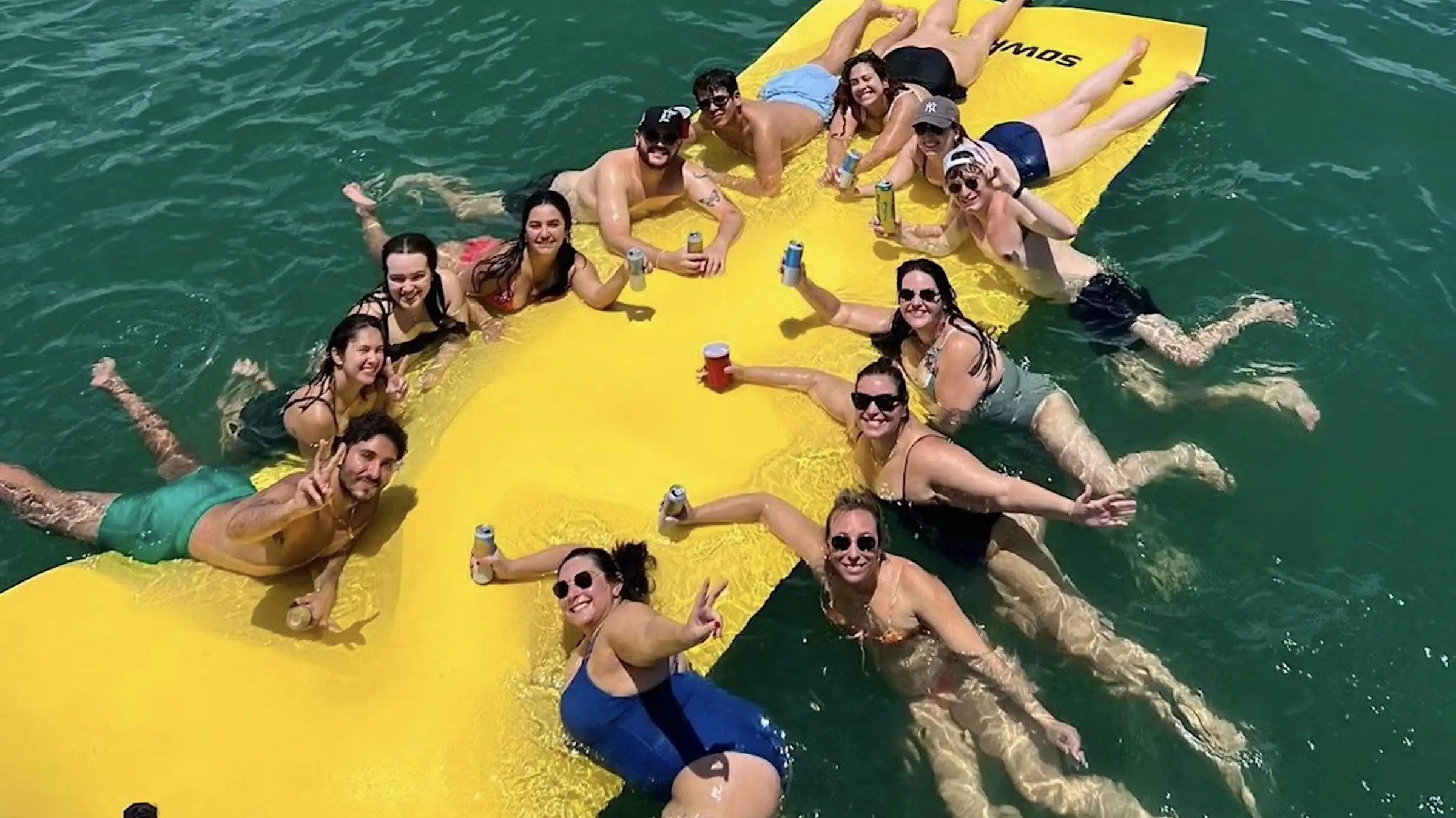 Group of people enjoying a sunny day on a yellow floating mat in the water, holding drinks and smiling.