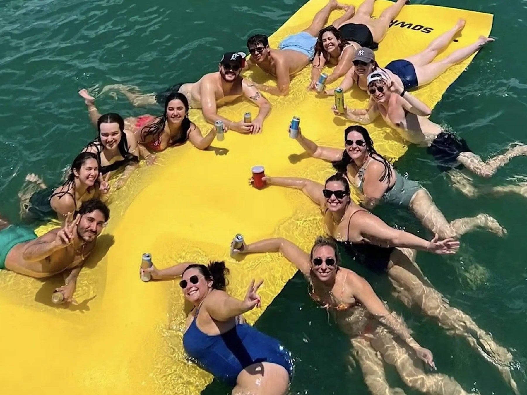 Group of people enjoying a sunny day on a yellow floating mat in the water, holding drinks and smiling.