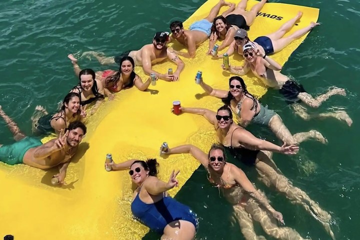 Group of people enjoying a sunny day on a yellow floating mat in the water, holding drinks and smiling.