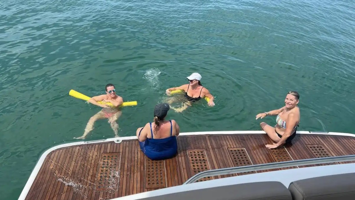 Four people enjoying water activities near a boat. Two are swimming with noodles, two are on the deck.
