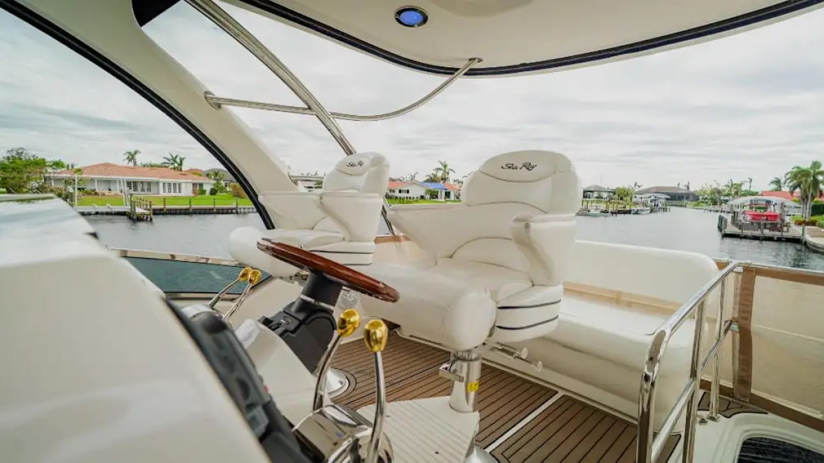 Boat helm with white seats and steering wheel, overlooking a calm canal with houses.