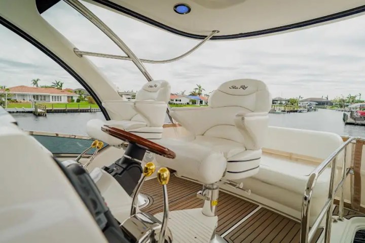 Boat helm with white seats and steering wheel, overlooking a calm canal with houses.