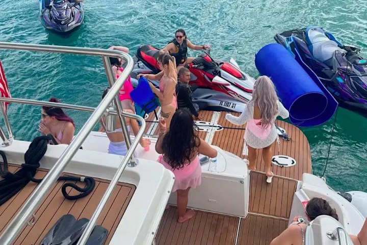 People on a yacht's deck with jet skis and a floating mat in the water.