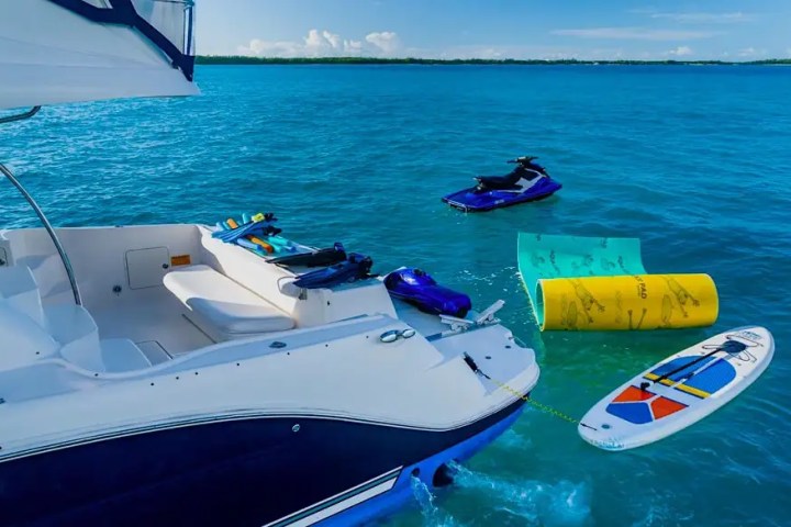 Boat with jet skis, paddleboard, and floating mat in blue ocean under clear sky.